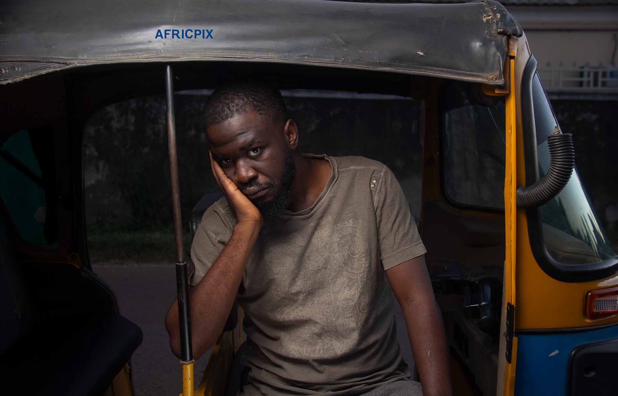 A sad and worried African man sitting in a tricycle with his hand on his cheek, looking worried.